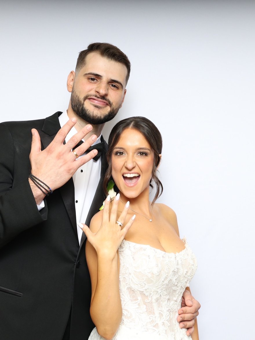 A smiling couple in a tuxedo and wedding dress holding up their hands to show their rings