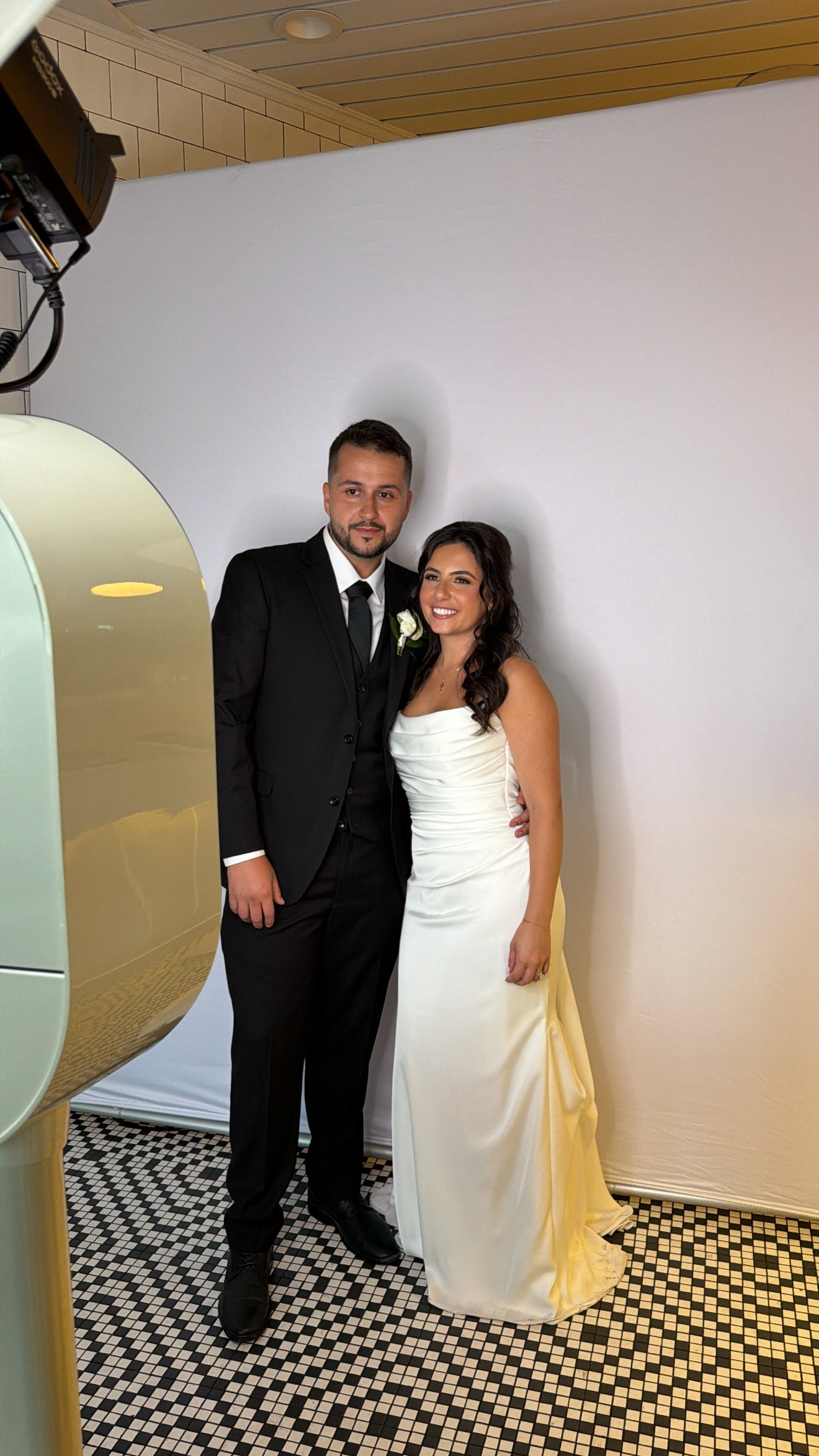 A couple in a black suit and white dress standing beside a white photo booth