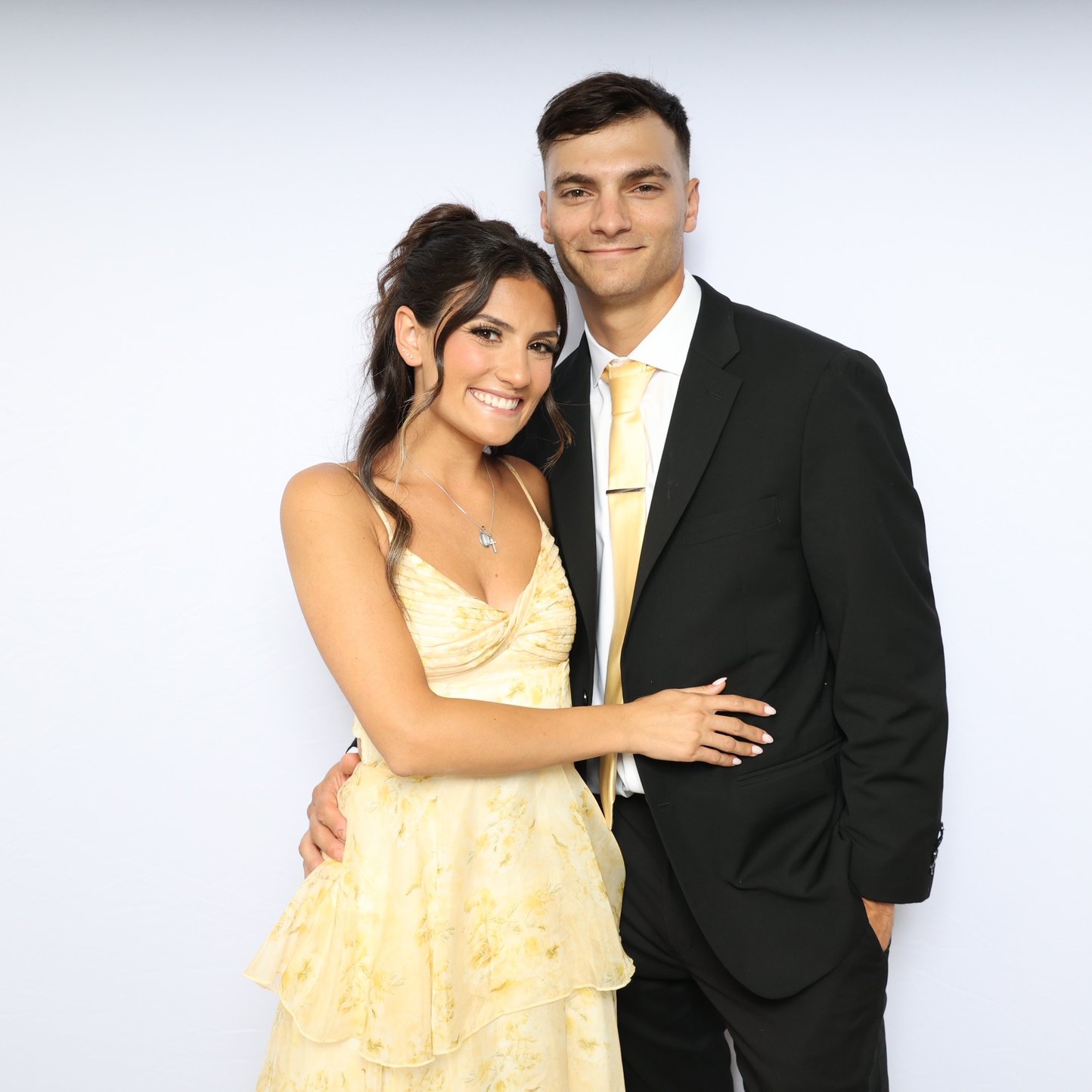 A couple in a yellow dress and black suit posing in front of a white backdrop