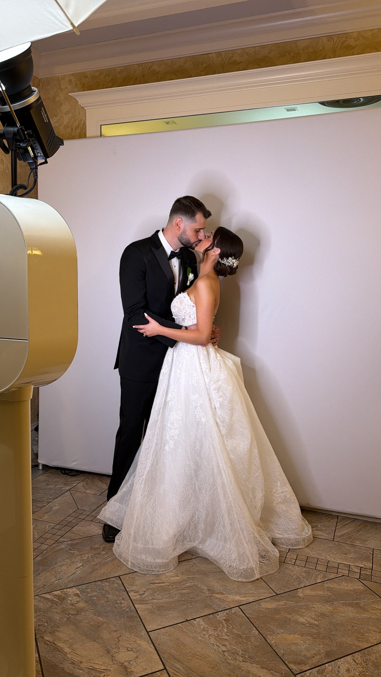 A couple in a tuxedo and wedding dress kissing beside a white photo booth